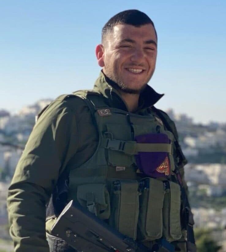 אלקנה מחייך, לבוש מדים ואפוד קרבי, ועל כתפו כומתה סגולה; ברקע עיר על גבעה