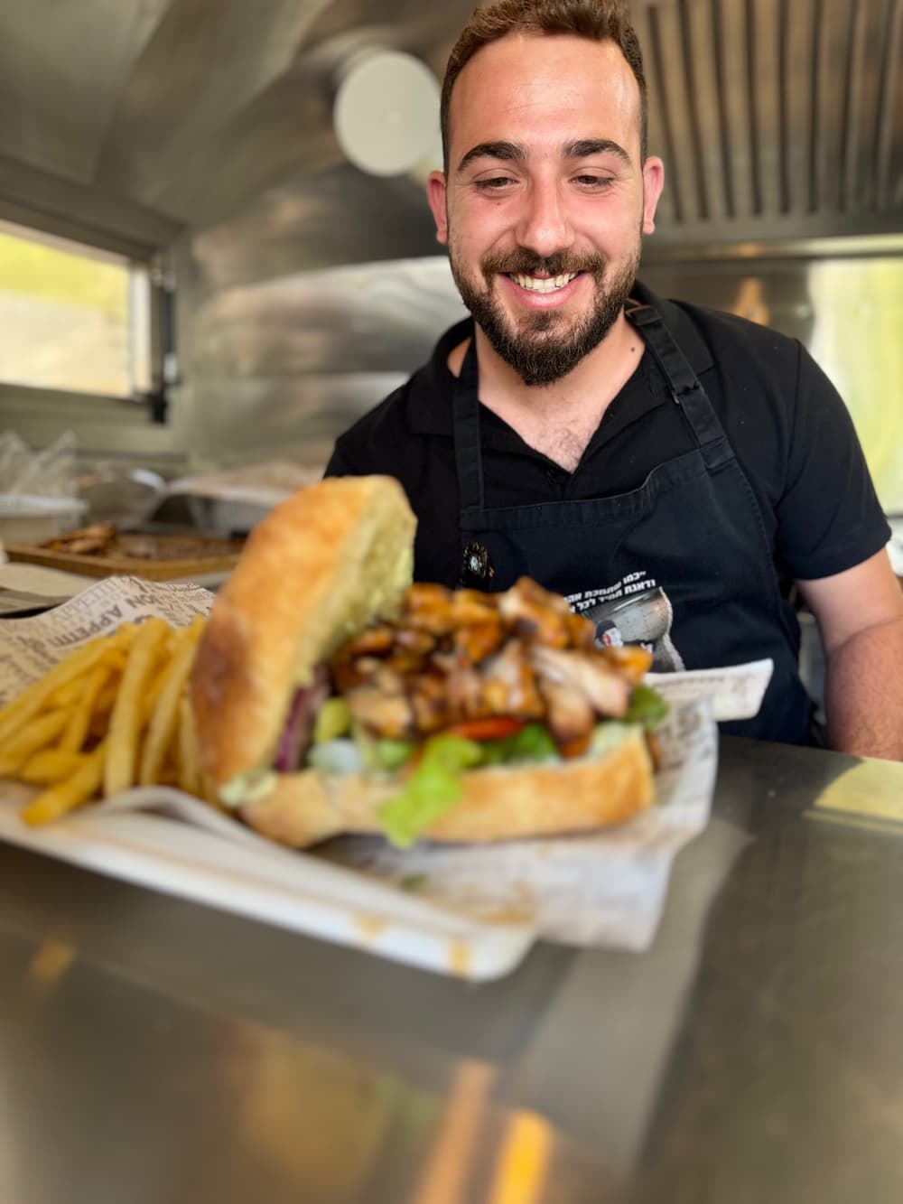 מתנדב מחייך בתוך הפודטראק ומגיש כריך גדול עם צ׳יפס