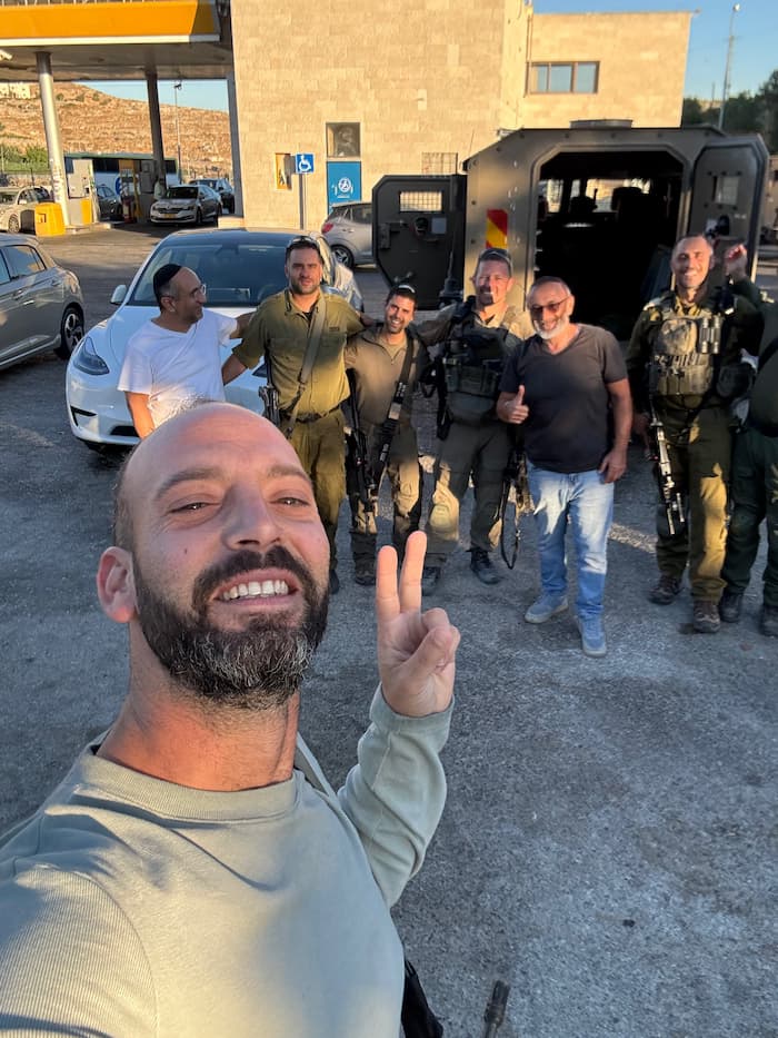 גבר מצלם סלפי ומסמן וי באצבעות; מאחוריו קבוצת אזרחים וחיילים ליד רכב צבאי משוריין