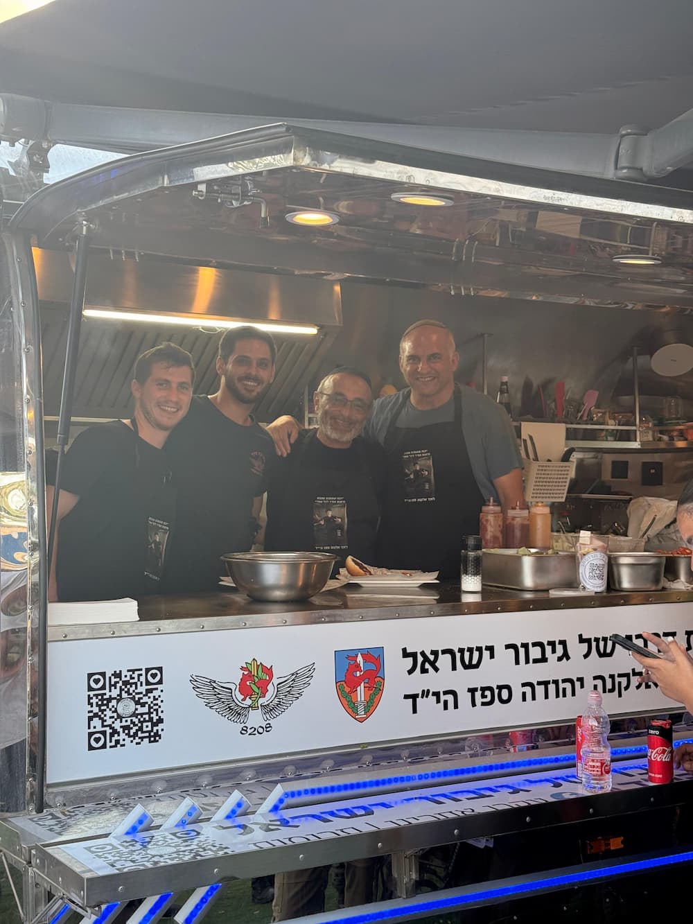 הפודטראק של יד אלקנה מואר בערב, וארבעה מתנדבים מחייכים מאחורי הדלפק. על הדלפק כתוב: גיבור ישראל אלקנה יהודה ספז הי״ד