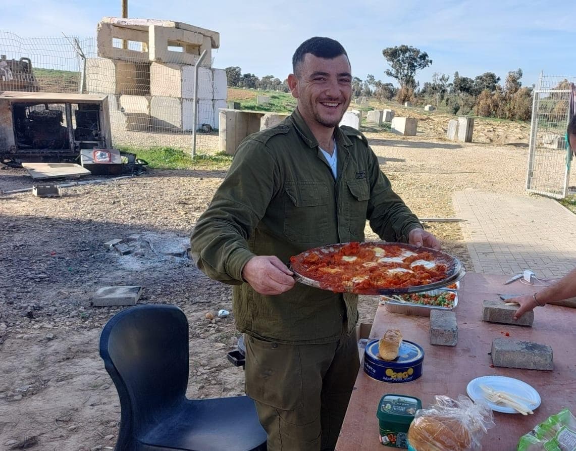אלקנה במדים מחייך ומגיש מחבת גדולה של שקשוקה ליד שולחן בשטח