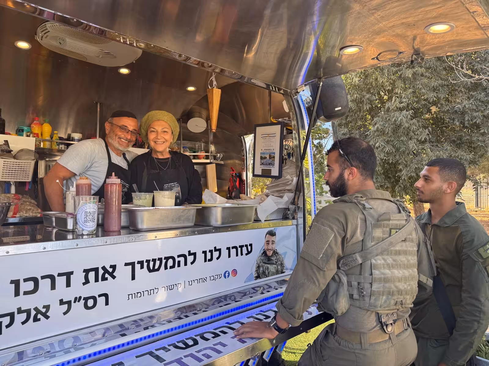 הפודטראק של יד אלקנה: גבר ואישה מחייכים מאחורי הדלפק, ושני חיילים עומדים מולם. על הדלפק שלט לזכר רס״ל אלקנה